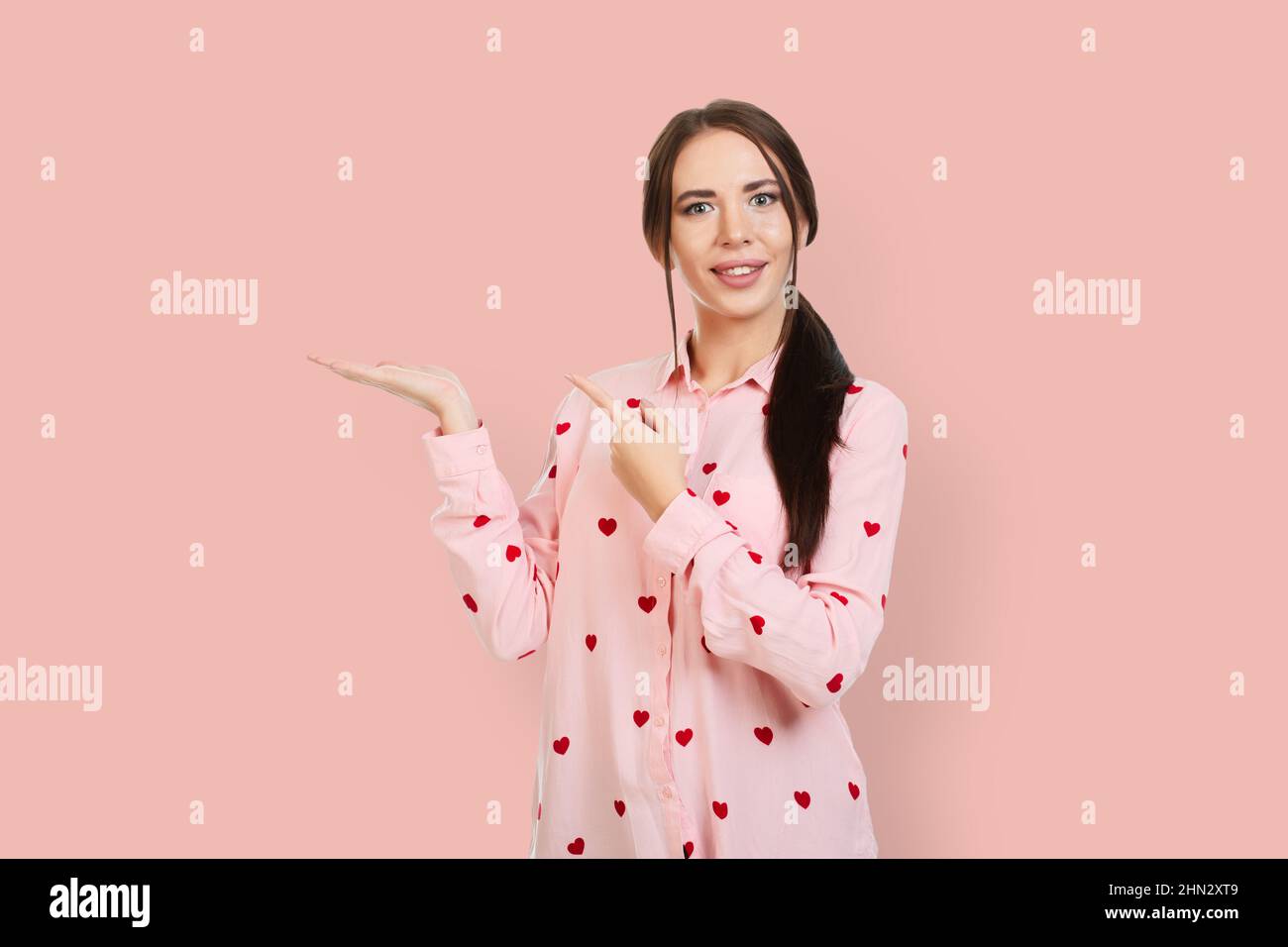 Giovane, bella ragazza con un sorriso, su sfondo rosa in una camicia rosa con  cuori rossi, punta le dita al posto per promozioni, offerte o pubblicità  Foto stock - Alamy