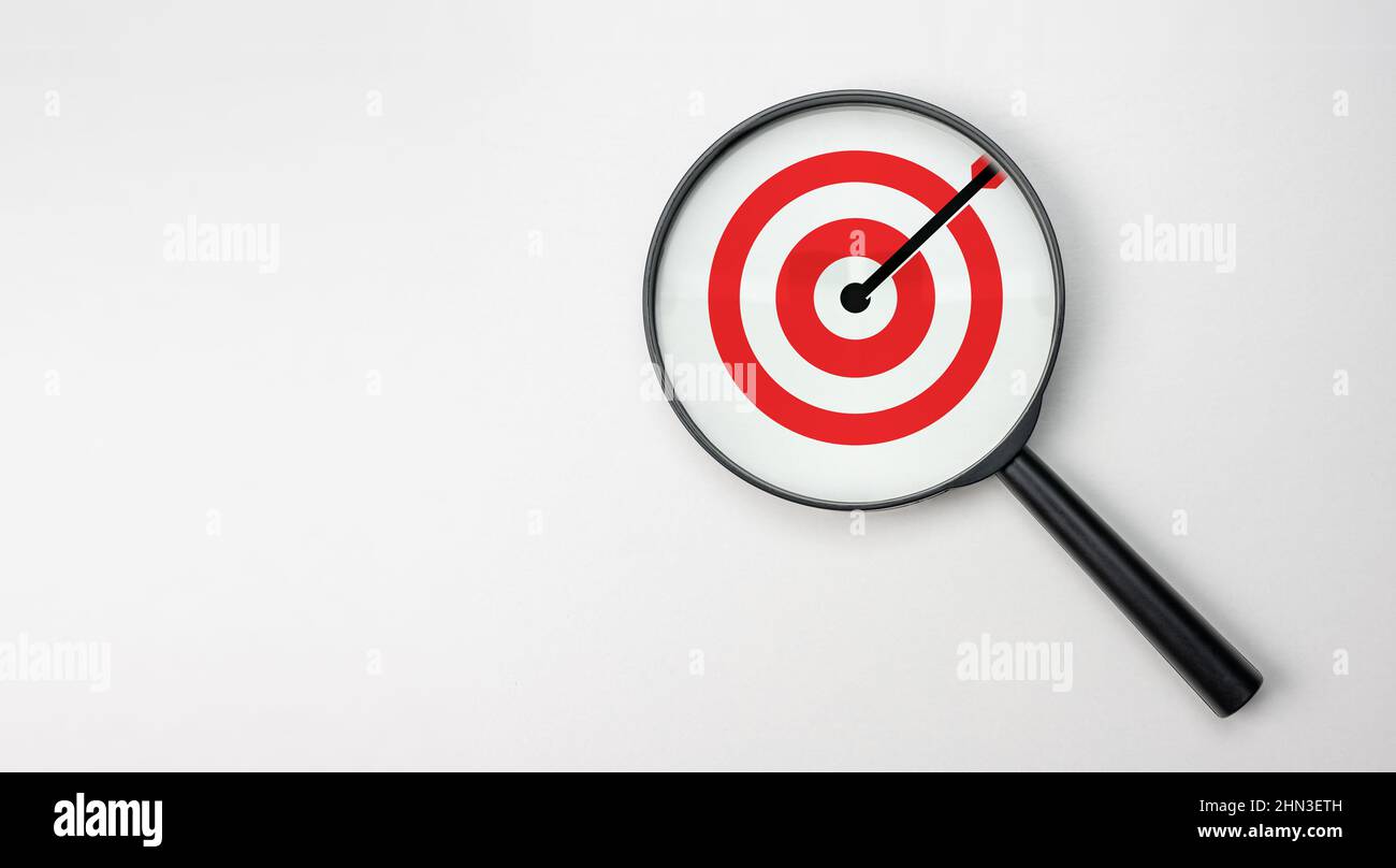 Target board all'interno della lente di ingrandimento per focalizzare l'obiettivo aziendale su sfondo bianco e spazio di copia. Ricerca e analisi dei target. Mano con magn Foto Stock