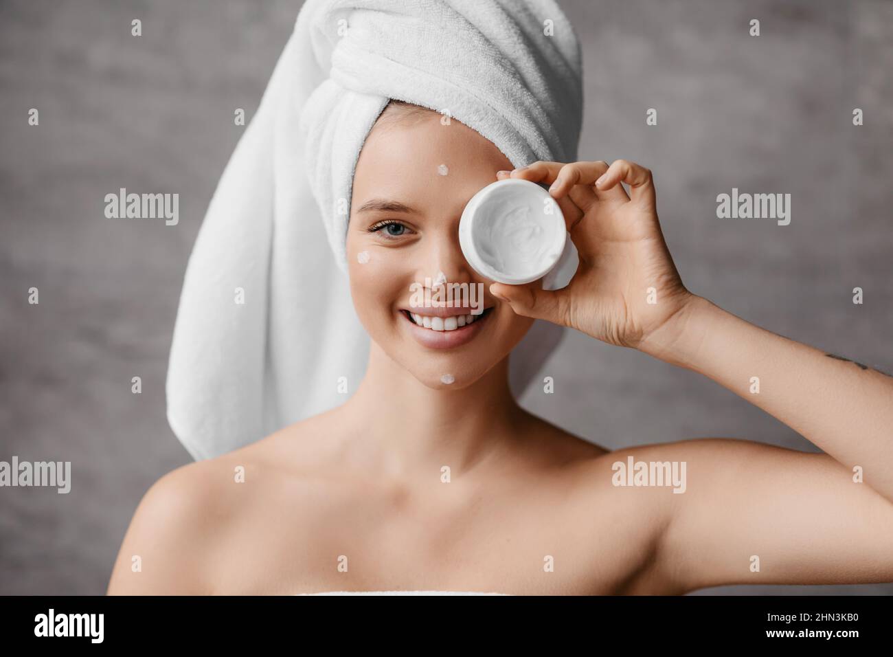 Pelle idratante dopo la doccia. Bella donna millenaria in asciugamano che copre il suo occhio con vasetto di crema in bagno interno Foto Stock
