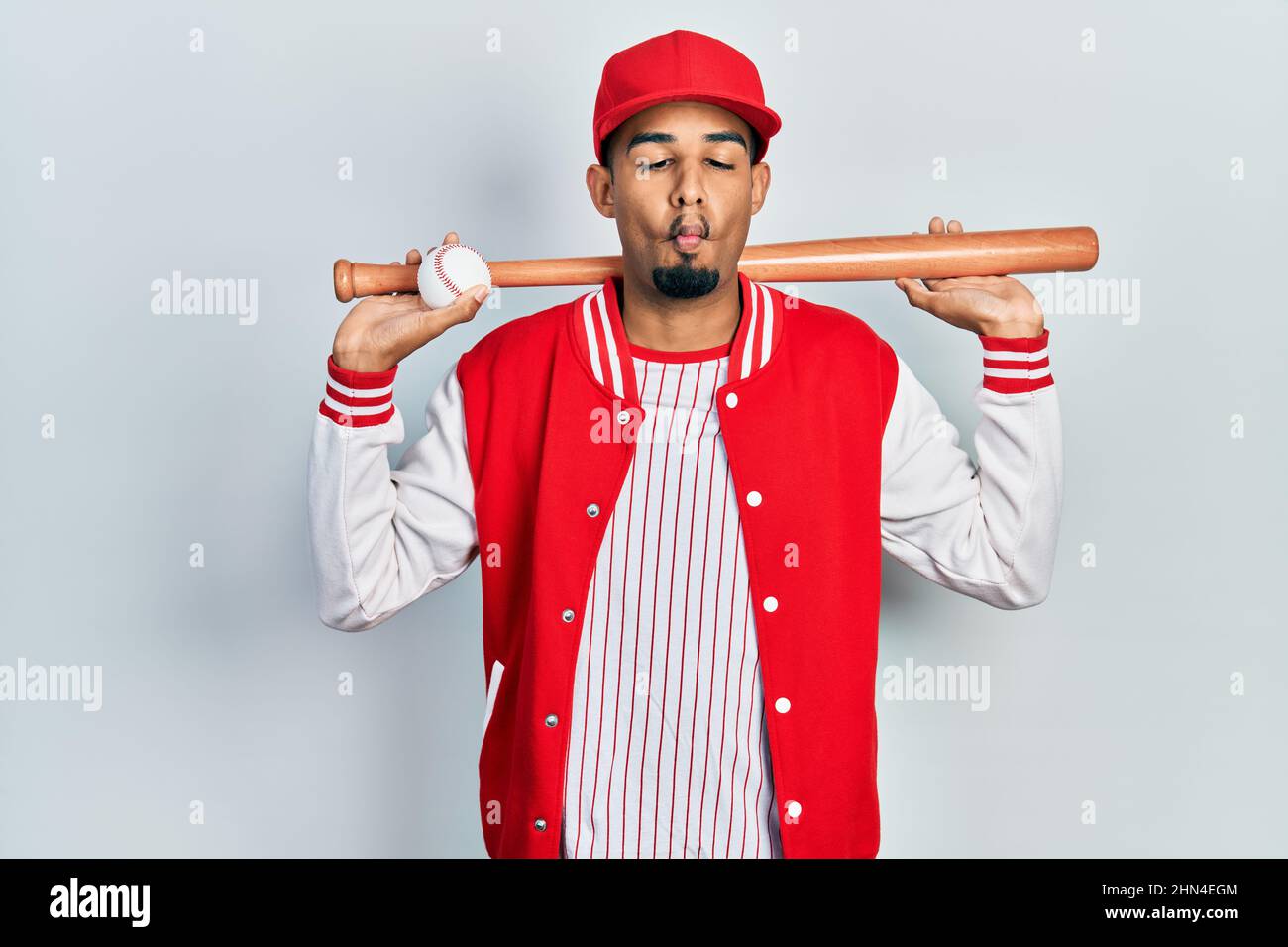 Giovane afroamericano che gioca a baseball tenendo la mazza e la palla facendo il pesce faccia con la bocca e gli occhi squinting, pazzo e comico. Foto Stock