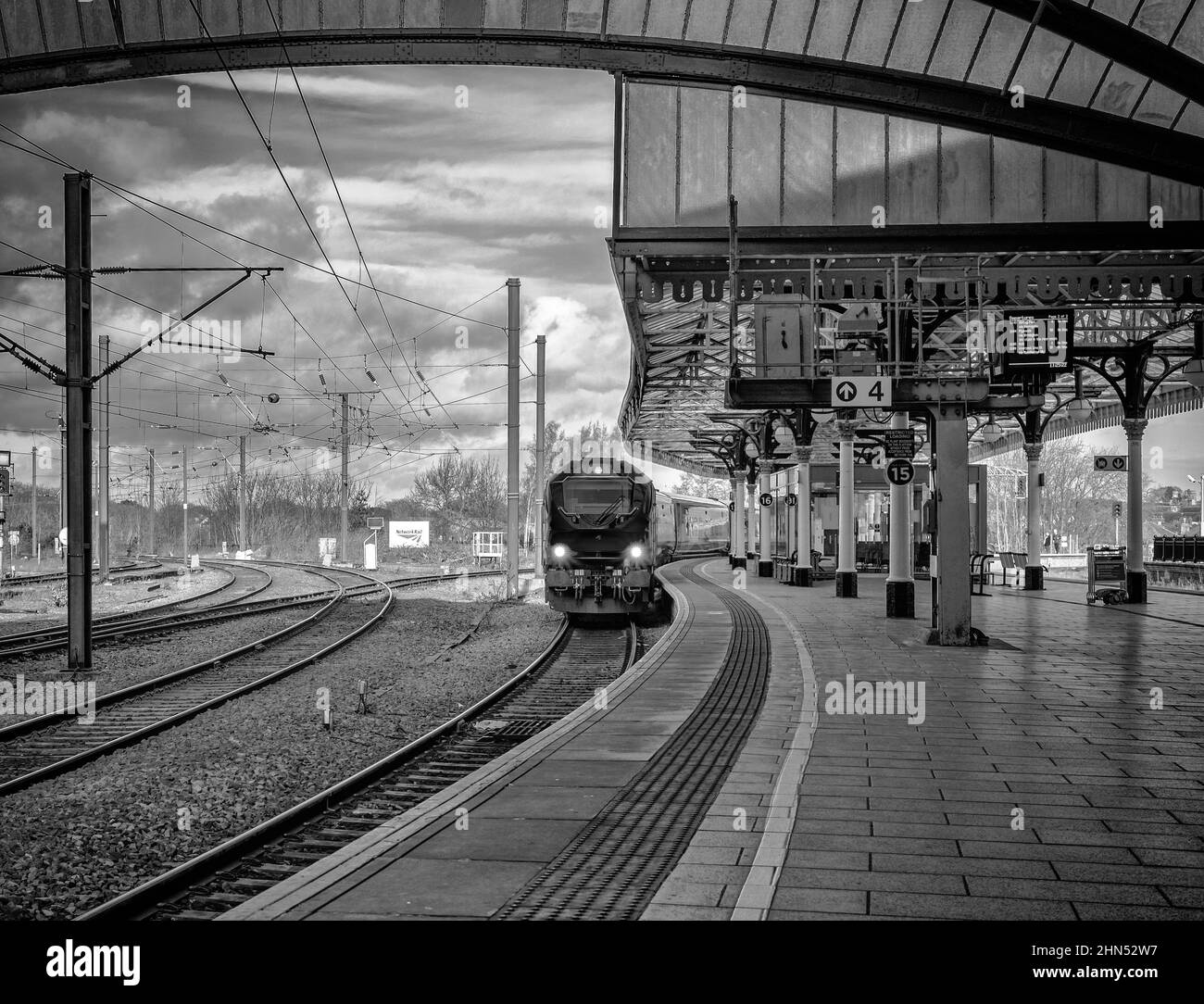 Un treno si avvicina ad una stazione accanto ad un baldacchino storico con colonne. Sopra si trova una tettoia ad arco di metallo e vetro. Foto Stock