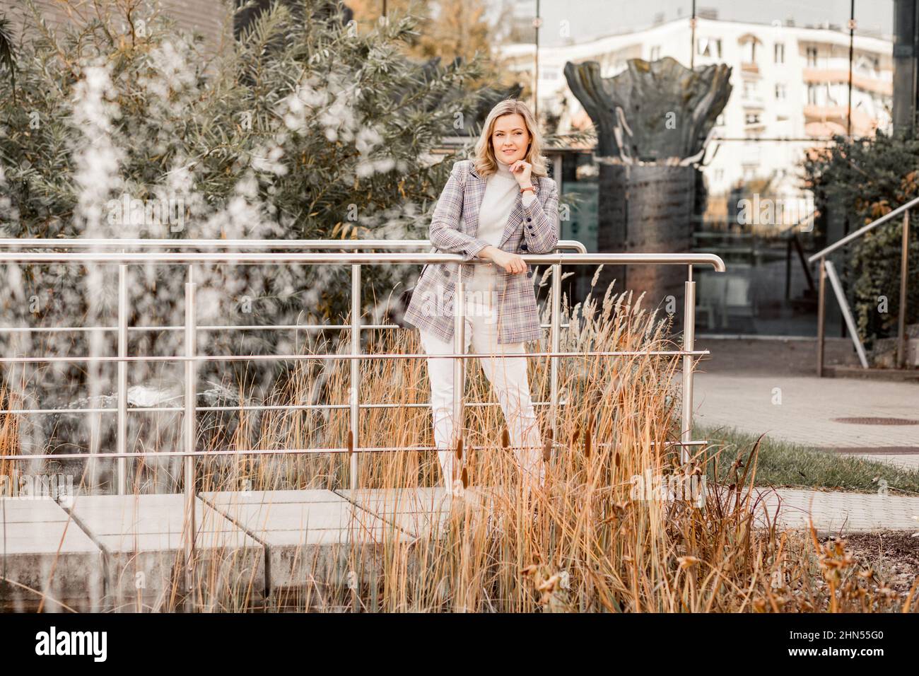 Bella donna sorridente di mezza età in giacca a scacchi e maglione grigio con girocollo poggiano il gomito sul ponte di metallo. Foto Stock