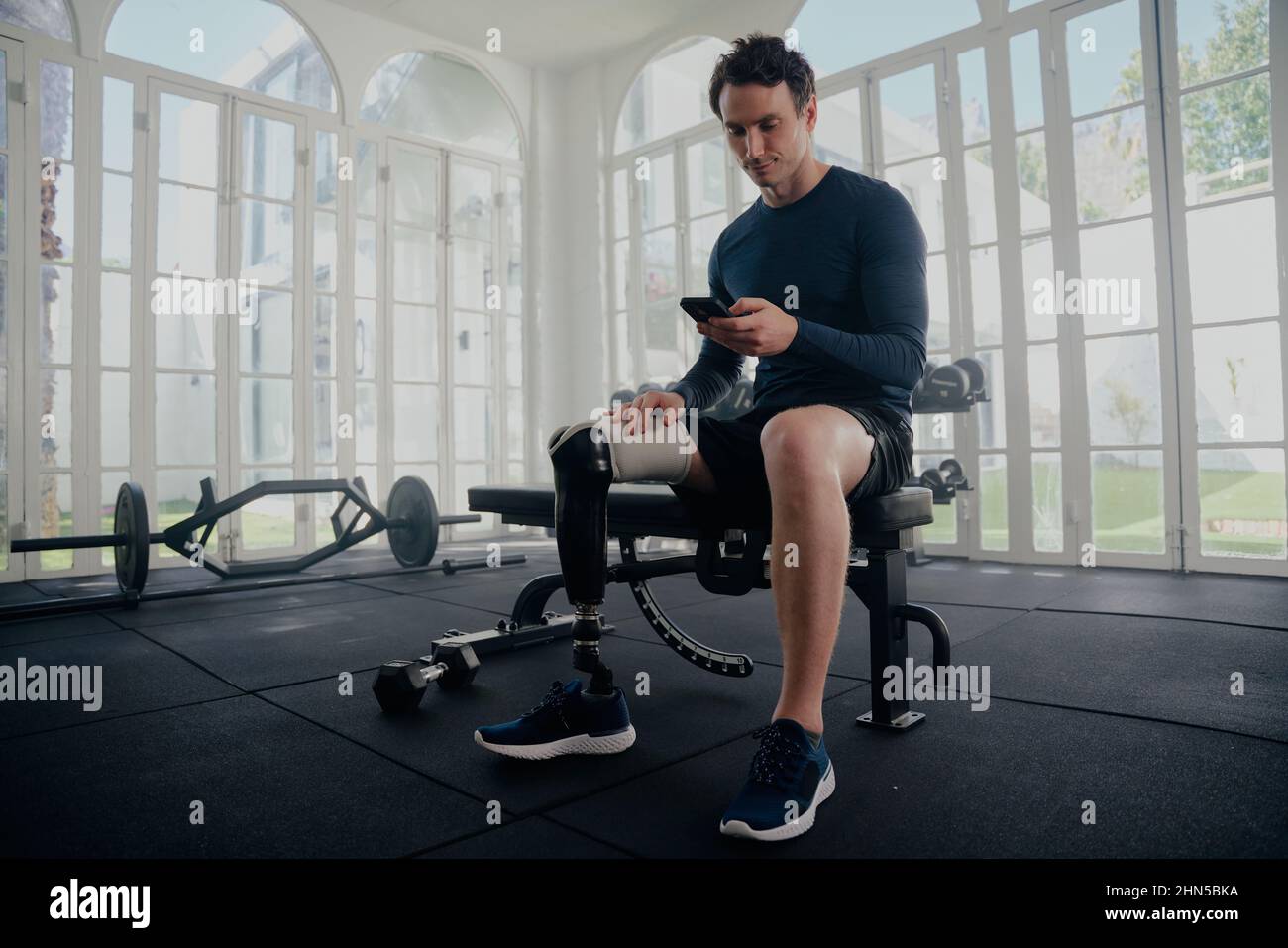 Atleta Paralimpico sul suo mobile in palestra di allenamento. Uomo con protesi seduta su panca guardando il telefono cellulare . Foto Stock