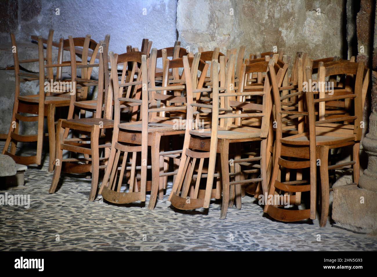 Sedie in legno impilate in un ambiente rustico che mostra artigianato e storia vicino a un pavimento di ciottoli in un ambiente scarsamente illuminato. Francia Foto Stock