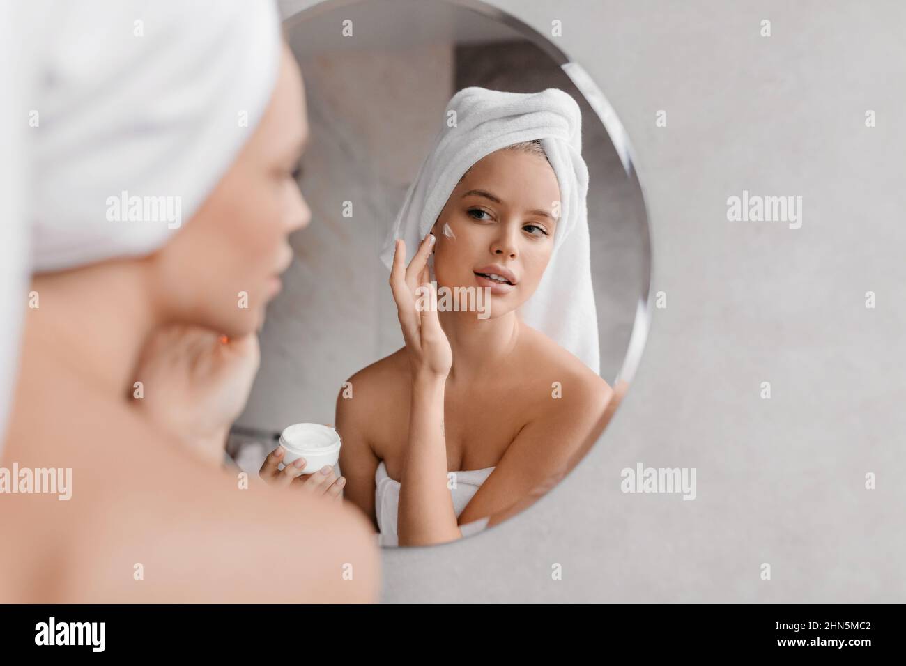 Routine di bellezza domestica. Giovane donna che applica crema idratante sul viso guardando mirore in bagno, spazio libero Foto Stock