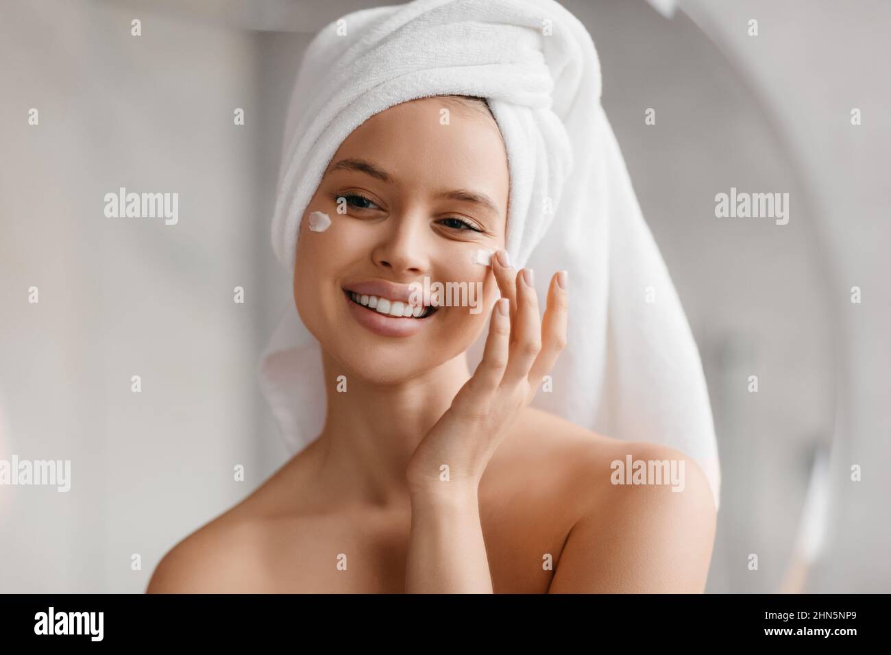 Colpo di testa della donna che applica la crema facciale sulle guance, sorridendo al suo riflesso nello specchio, godendo la routine di bellezza Foto Stock