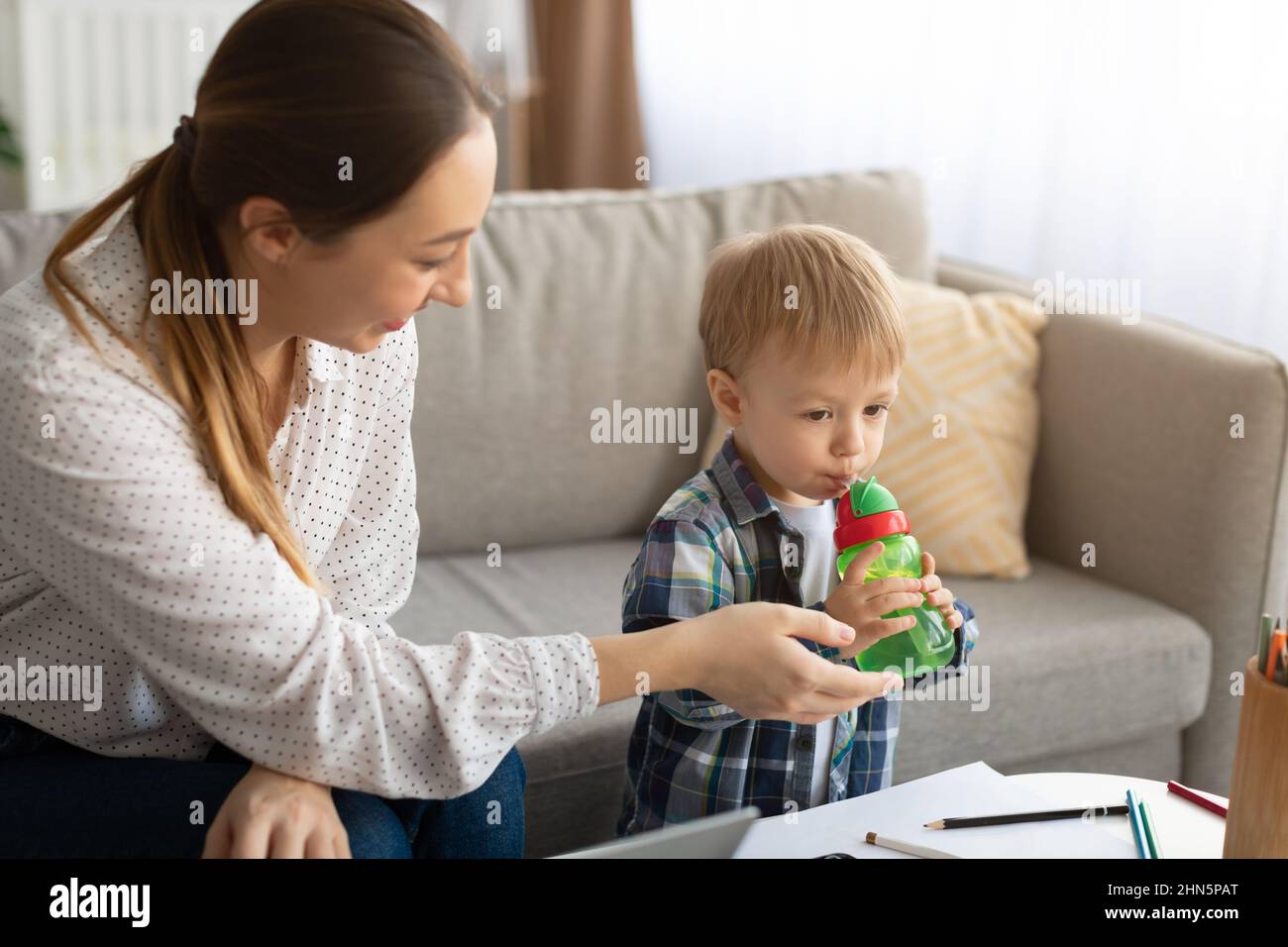 Concetto di congedo di maternità. Mamma giovane che dà la bottiglia con acqua a suo figlio mentre lavora in linea da casa Foto Stock