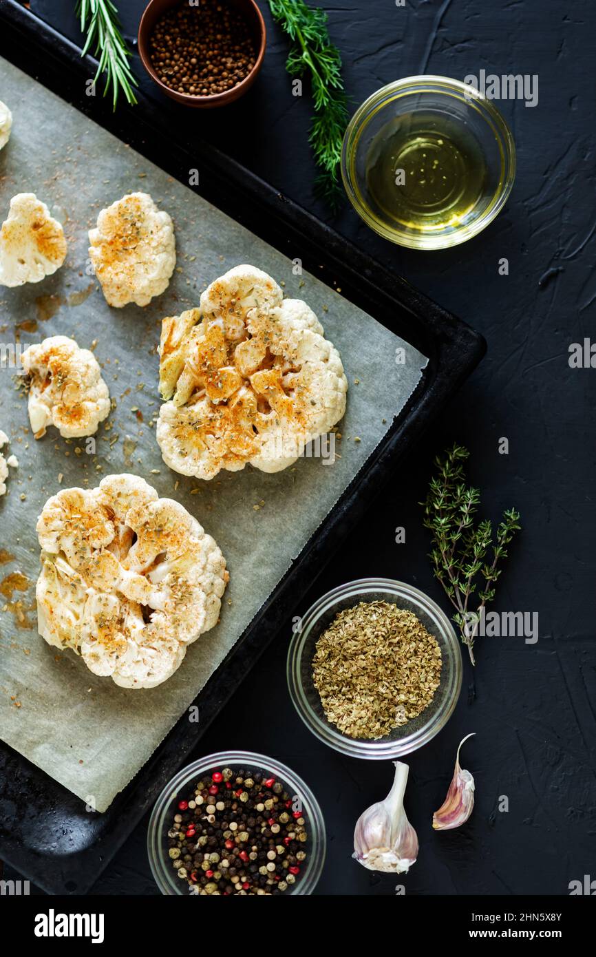 Cucina con bistecca di cavolfiore. Il cavolfiore crudo cosparso di spezie si trova su una teglia da forno. Olio d'oliva, erbe, varie spezie nelle vicinanze. Sfondo scuro. Foto Stock