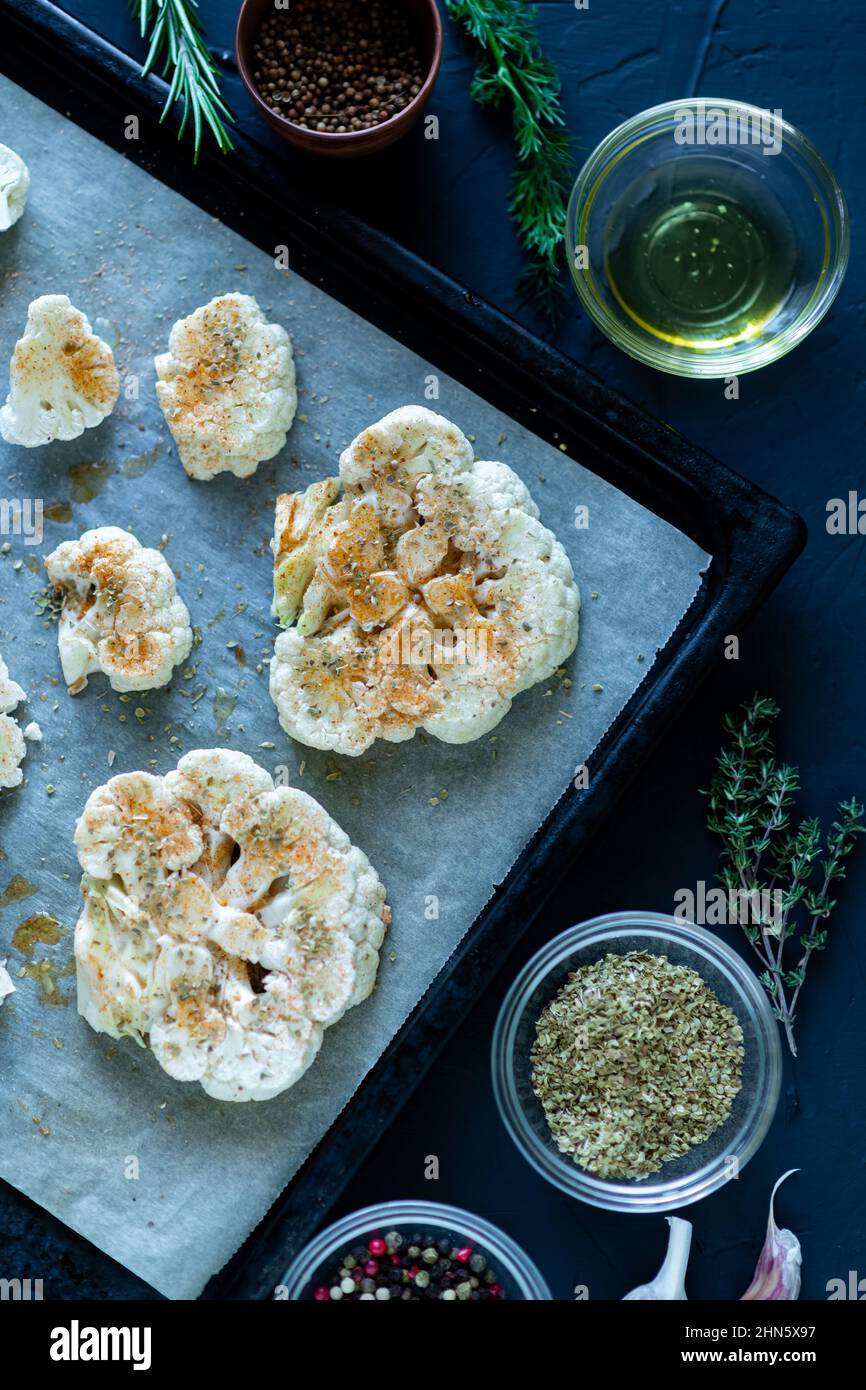 Cucina con bistecca di cavolfiore. Il cavolfiore crudo cosparso di spezie si trova su una teglia da forno. Olio d'oliva, erbe, varie spezie nelle vicinanze. Sfondo scuro. Stagno Foto Stock