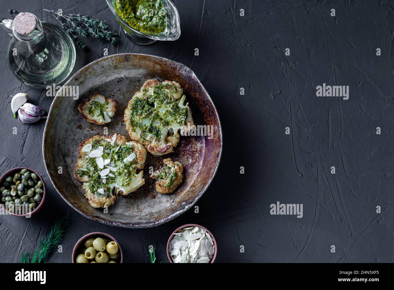 Bistecca di cavolfiore con spezie, salsa di chimichurri, fiocchi di mandorle. Capperi, olive, erbe, fianco a fianco. Sfondo scuro. Cibo vegetariano. CopySpace. Stagno Foto Stock