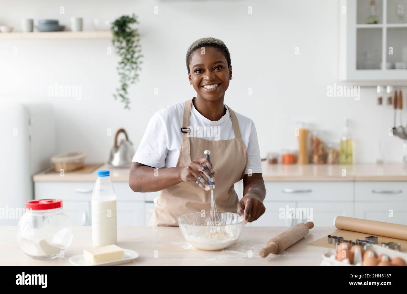 Sorridente chef donna afroamericana piuttosto millennial in grembiule facendo pasta in interni di cucina minimalista chiaro Foto Stock