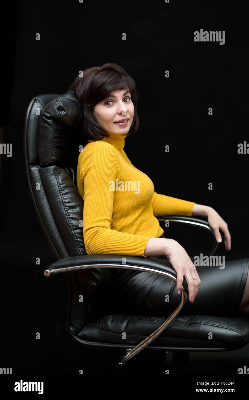 Una donna brunetta di 35-40 anni con un sorriso è seduta in un enorme sedia in pelle sfondo nero. Foto Stock