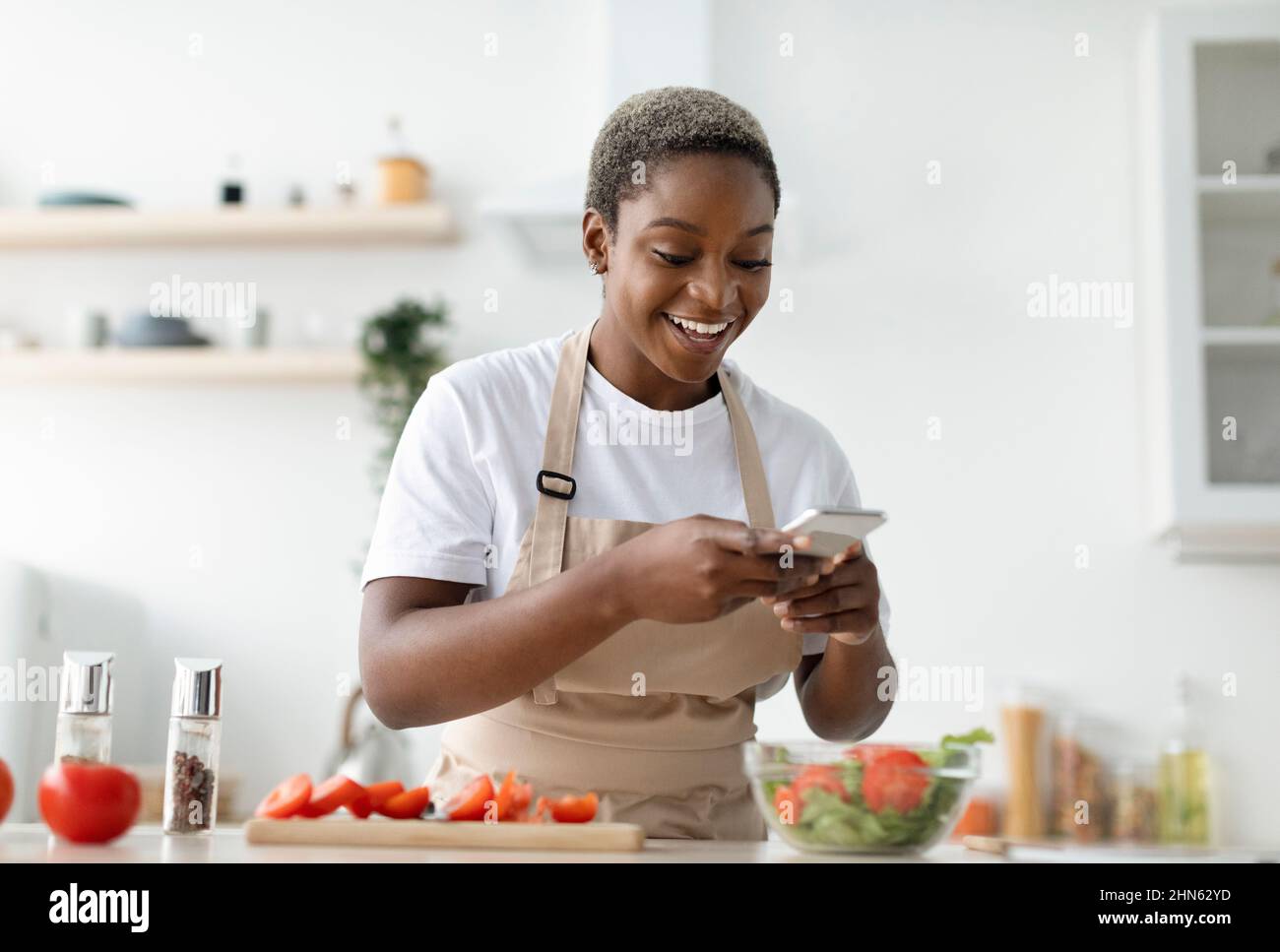 Sorridente giovane ragazza afroamericana graziosa blogger in grembiule facendo foto di insalata per il blog di cibo Foto Stock