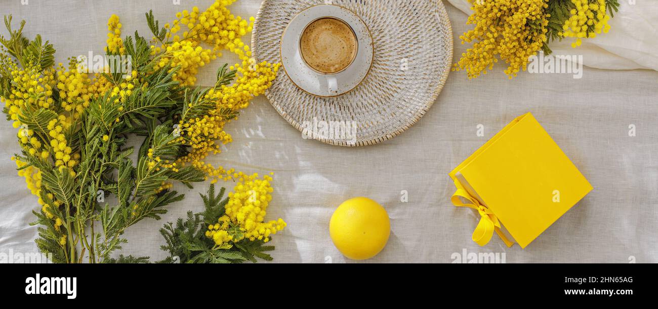 Rami di fiori gialli di mimosa su lenzuola di lino bianco, tazza o caffè, macaroon gialli francesi, pompelmo e confezione regalo gialla. Saluto per Inte Foto Stock