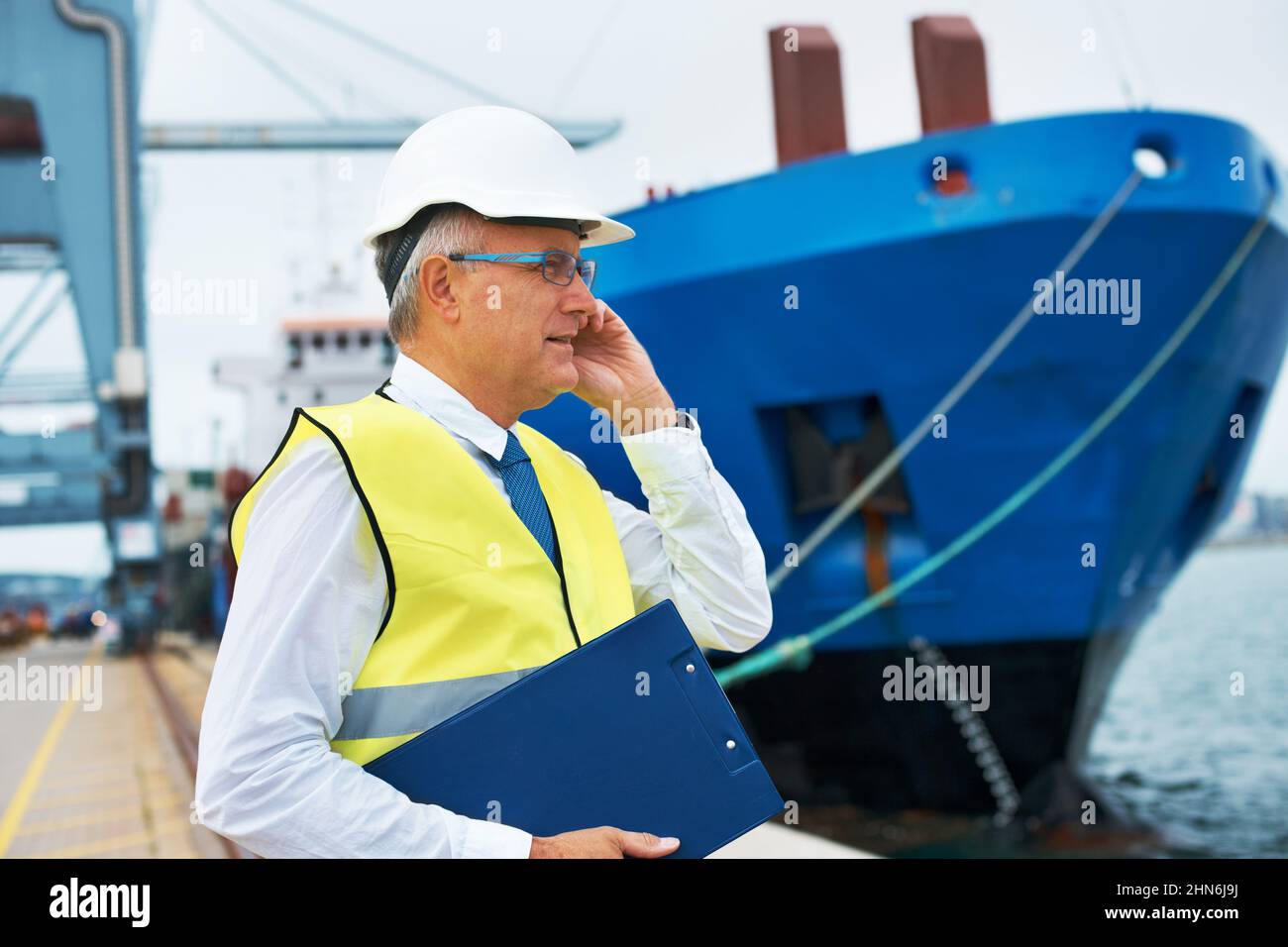 Dove è il mio cliente. Un lavoratore portuale in piedi al porto durante l'attività dell'industria marittima mentre prende una chiamata sul suo mobile. Foto Stock