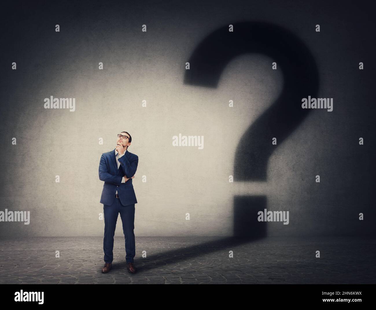 Un uomo d'affari pensivo che getta un'ombra di segno di interrogatorio sul muro. Domande aziendali e concetto decisionale. Pensare e trovare soluzioni Foto Stock