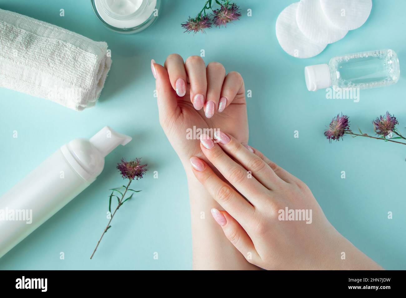 Belle mani femminili e prodotti cosmetici su sfondo blu. Bottiglia di crema, batuffoli di cotone e fiori. Cura del corpo, concetto di benessere. Foto Stock