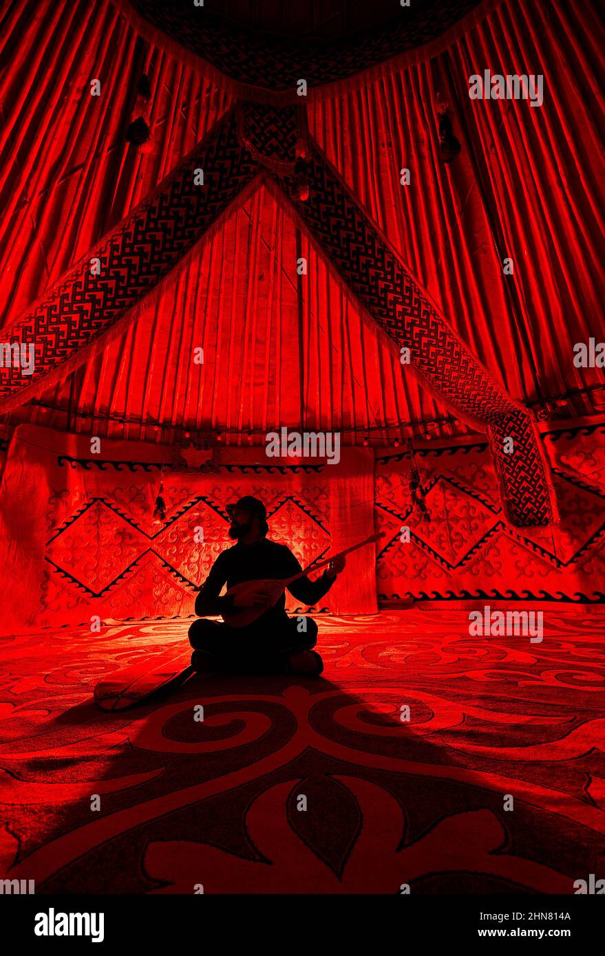 Silhouette dell'uomo che tiene e gioca lo strumento di corda del dombra all'interno della casa nomade di Yurt contro il tappeto con motivi etnici illuminati da luce rossa interna Foto Stock