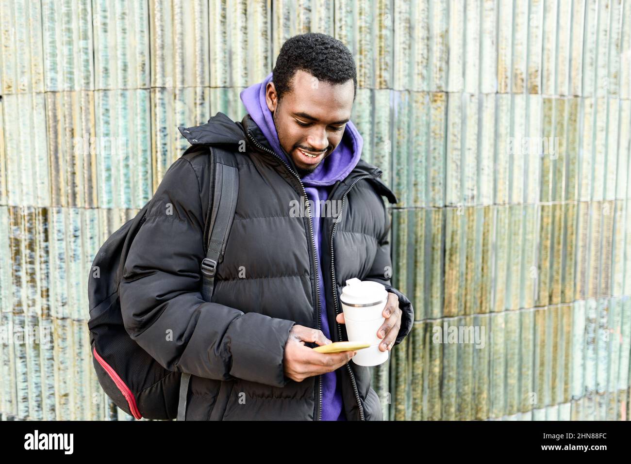 Uomo afro-americano positivo in outerwear con zero rifiuti takeaway tazza di bevanda calda. Sorride e naviga sui social media sul cellulare mentre si trova vicino a un muro traballante sulla strada della città Foto Stock