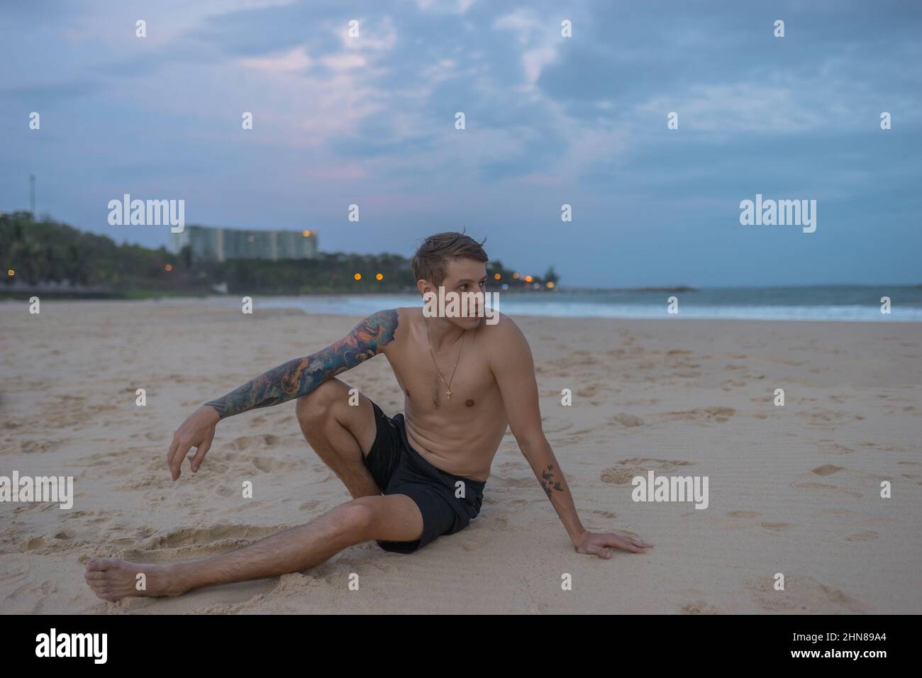 Ritratto a tutta lunghezza di bel caucasico seduto sulla sabbia sulla spiaggia al tramonto, tatuaggio a braccio pieno. Foto di alta qualità Foto Stock