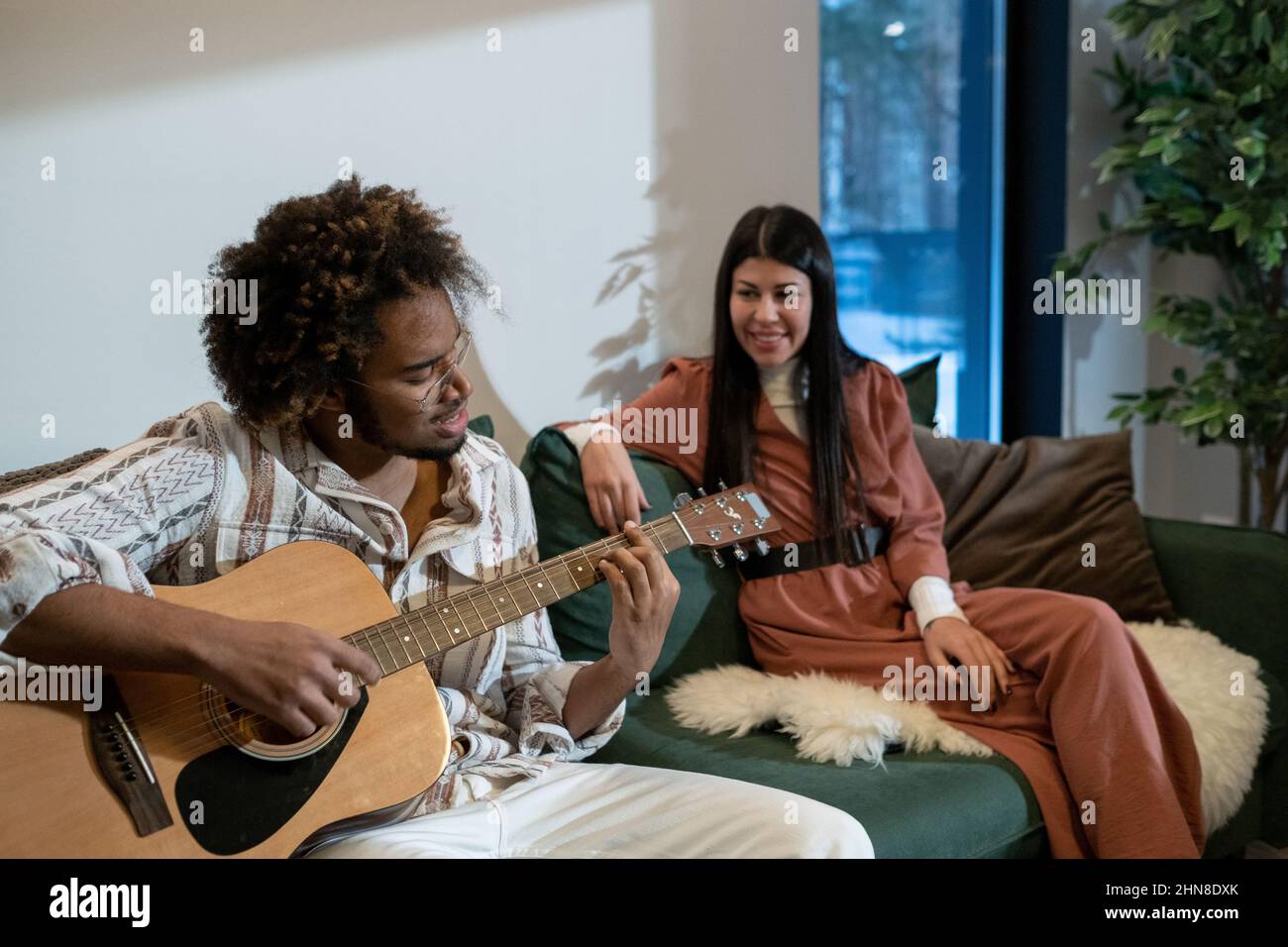 Giovane africano che suona la chitarra e canta una canzone mentre si siede sul divano insieme alla sua ragazza nella stanza Foto Stock