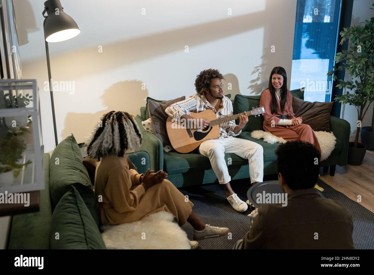 Giovane africano che suona la chitarra seduto sul divano con i suoi amici che cantano durante le loro vacanze in casa Foto Stock