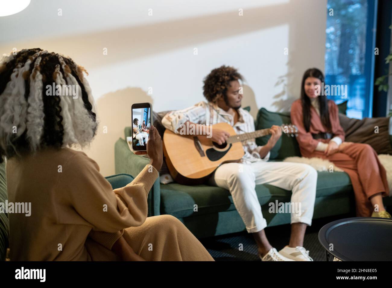 Vista posteriore della donna africana che fa il video del suo telefono cellulare di un uomo che suona la chitarra durante la loro festa domestica Foto Stock