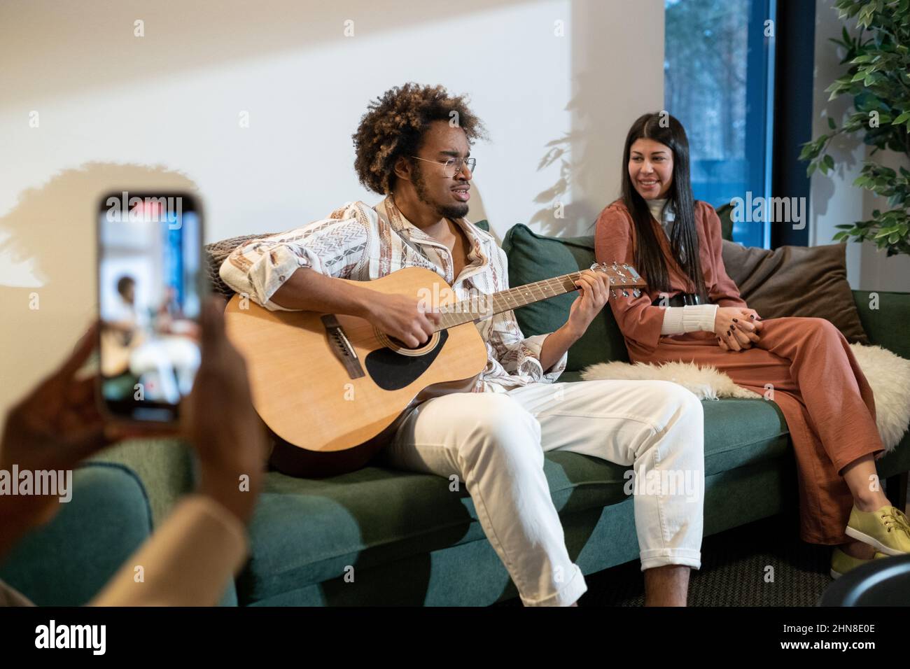Giovane africano che suona la chitarra per i suoi amici seduti sul divano in salotto mentre la donna fa una foto sul suo cellulare Foto Stock