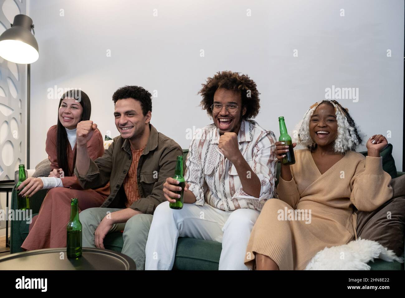 I giovani eccitati si siedono sul divano e guardano le partite sportive mentre bevono birra durante le feste di casa Foto Stock