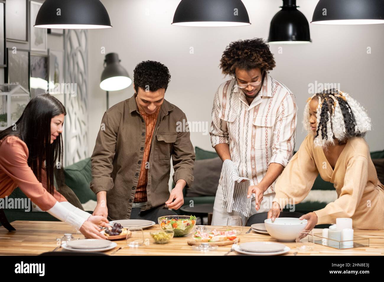 Gruppo di giovani amici che mettono insieme il tavolo da pranzo mettendo il pasto e i piatti, si preparano per la cena partito Foto Stock