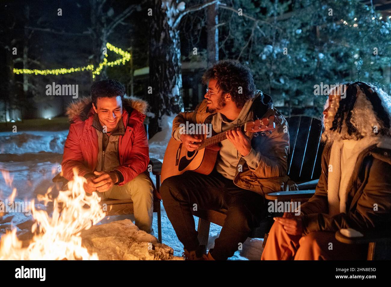 Giovane africano che suona la chitarra mentre si siede all'aperto vicino al fuoco con i suoi amici durante le vacanze invernali Foto Stock