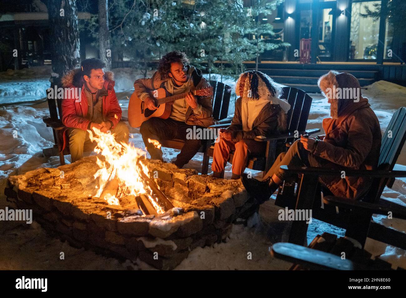 Gruppo di amici che suonano la chitarra e cantano insieme mentre si siedono vicino al fuoco all'aperto nella sera d'inverno Foto Stock