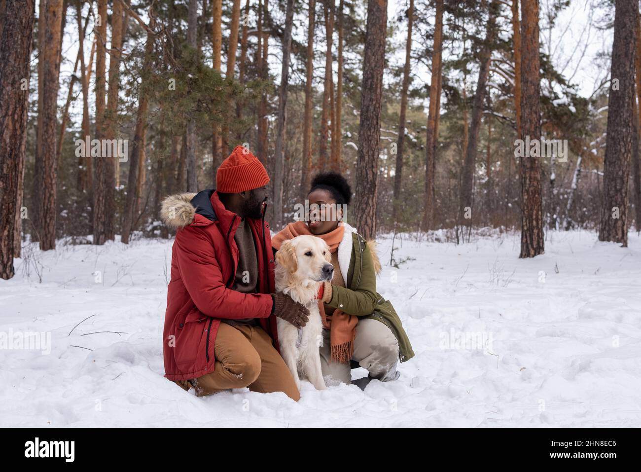 Coppia felice Africana che si parla l'un l'altro mentre si riposa sulla neve e abbraccia il loro cane durante la passeggiata nella foresta invernale Foto Stock