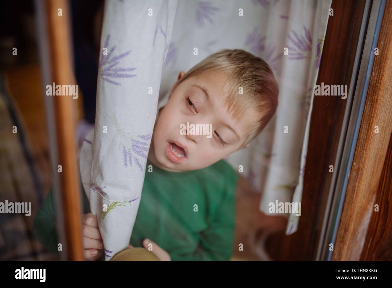 Ragazzino con sindrome di Down che guarda attraverso la finestra a casa. Foto Stock