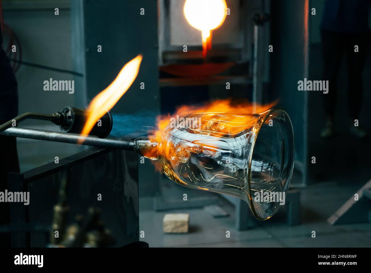 Primo piano di un artigiano soffiatore di vetro che forma il vetro fuso caldo a fuoco forte all'interno di un'officina. Lavorazione manuale del vetro da parte degli artigiani all'interno di un gla Foto Stock