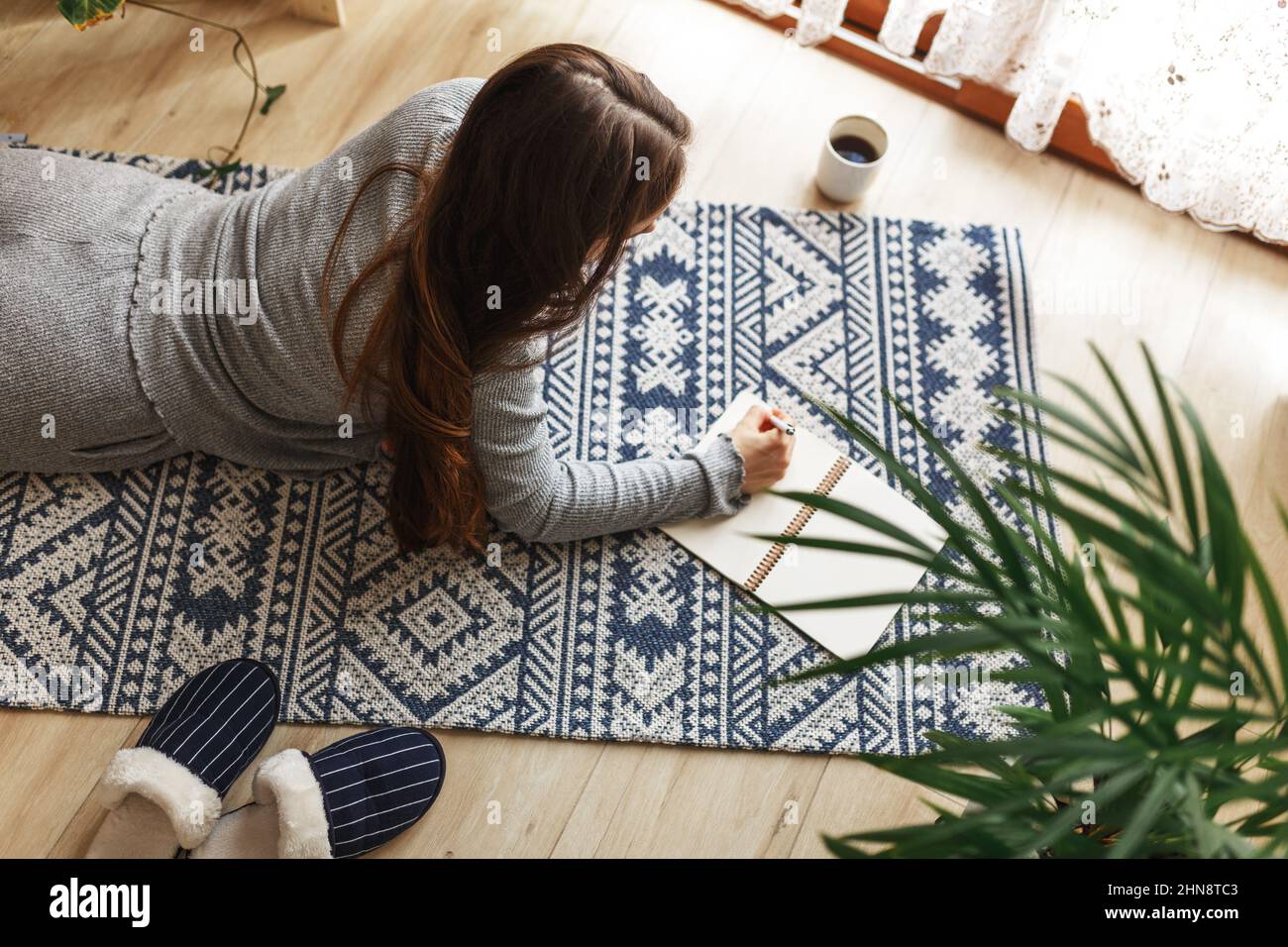 Giovane donna che scrive le pagine del mattino in diario, progettando la sua attività al mattino con il notebook e la penna nelle sue mani. Bere caffè interni casa moderna e luminosa sullo sfondo. Foto Stock