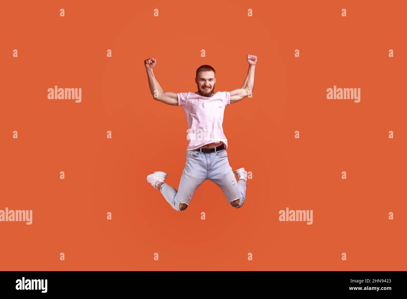 Ritratto di felice attraente uomo bearded che salta in aria le braccia sollevate, guardando la macchina fotografica, spazio di copia per l'annuncio, indossando la T-shirt rosa e jeans. Studio interno girato isolato su sfondo arancione. Foto Stock
