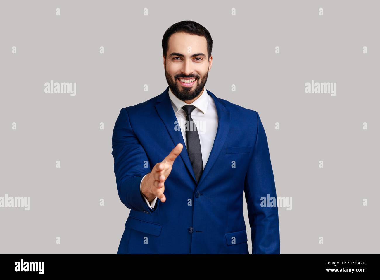 L'uomo positivo bearded che dà la mano al handshake e sorridente amichevole, consulente o responsabile di HR che offre lavoro, la collaborazione, indossando il vestito ufficiale di stile. Studio interno girato isolato su sfondo grigio. Foto Stock