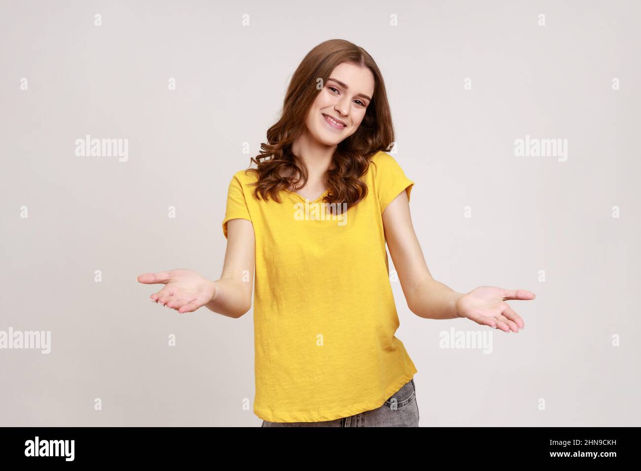 Si prega di prendere gratuitamente. Donna giovane e gentile in T-shirt gialla che accoglie con le braccia aperte e sorride gentilmente, guardando la macchina fotografica. Studio interno girato isolato su sfondo grigio. Foto Stock