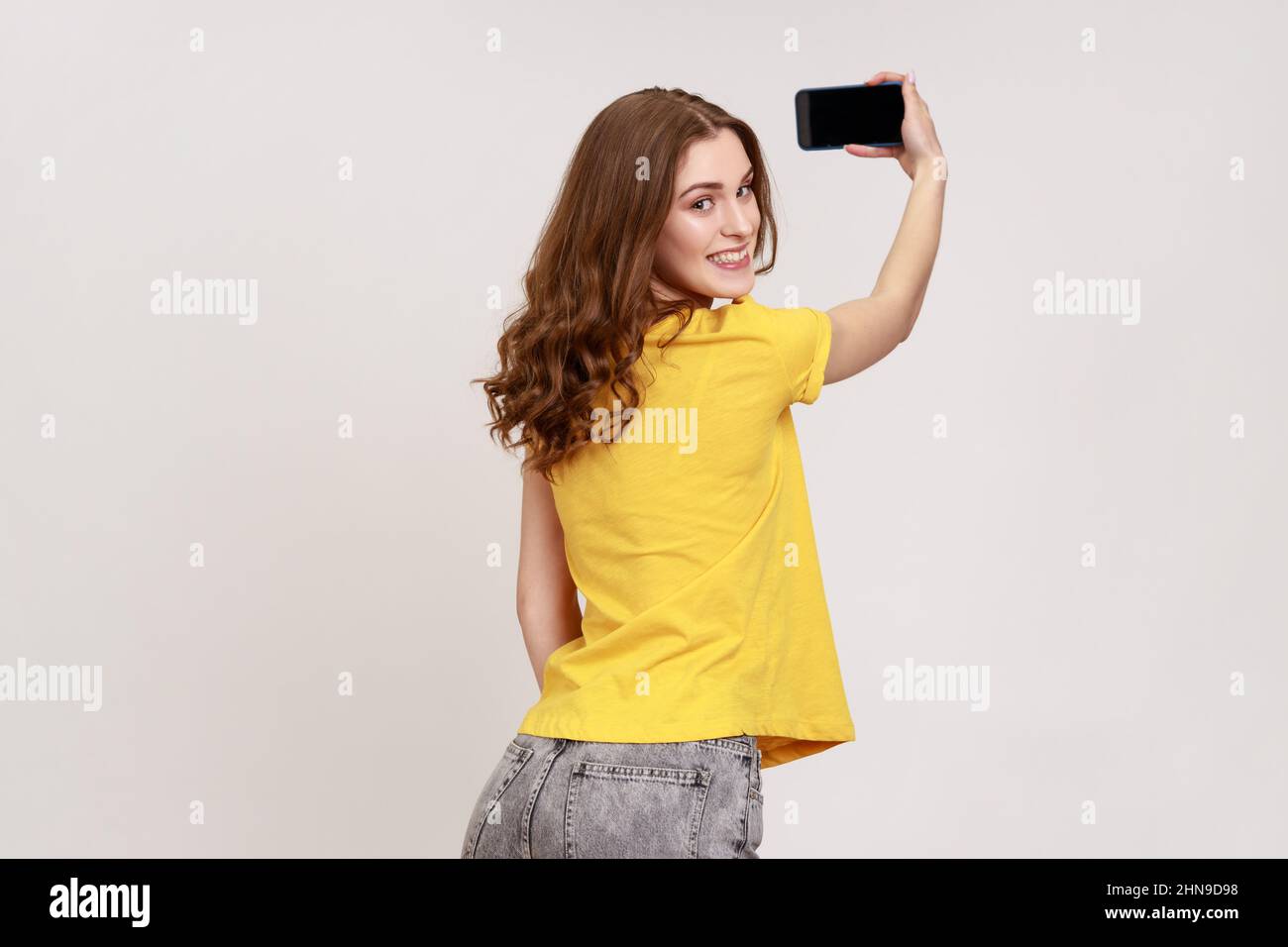 Vista posteriore ritratto di ragazza marrone capelli parlare videochiamata, prendere selfie sul telefono cellulare, mostrando display vuoto, guardando la fotocamera. Studio interno girato isolato su sfondo grigio. Foto Stock