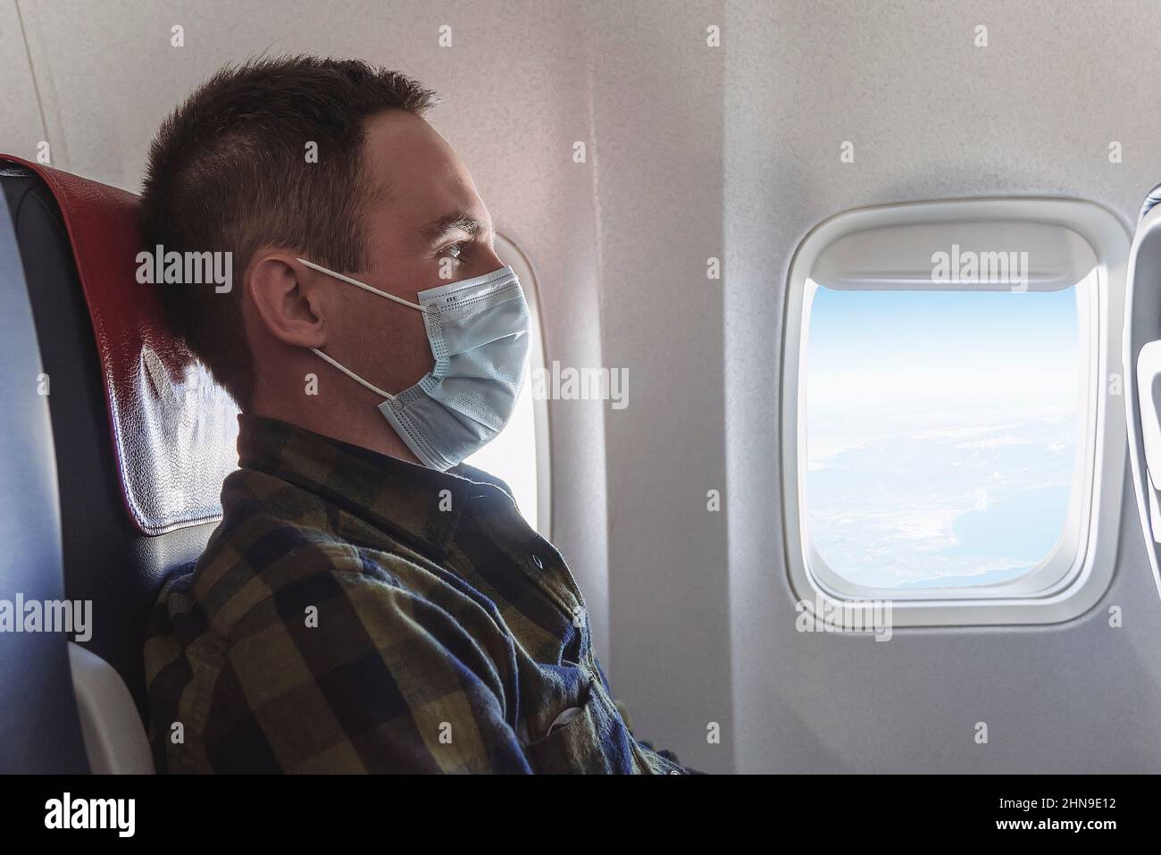 Un giovane uomo in una maschera medica blu siede nel sedile passeggero a bordo dell'aereo e guarda fuori la finestra. Volo sicuro durante lo scoppio del covi Foto Stock