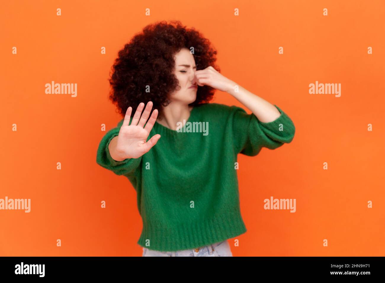 Donna con acconciatura afrofroche si macina con disgusto, trattenere il respiro, pizzicare il naso con le dita per evitare il cattivo odore e mostrare il gesto di arresto. Studio interno girato isolato su sfondo arancione. Foto Stock