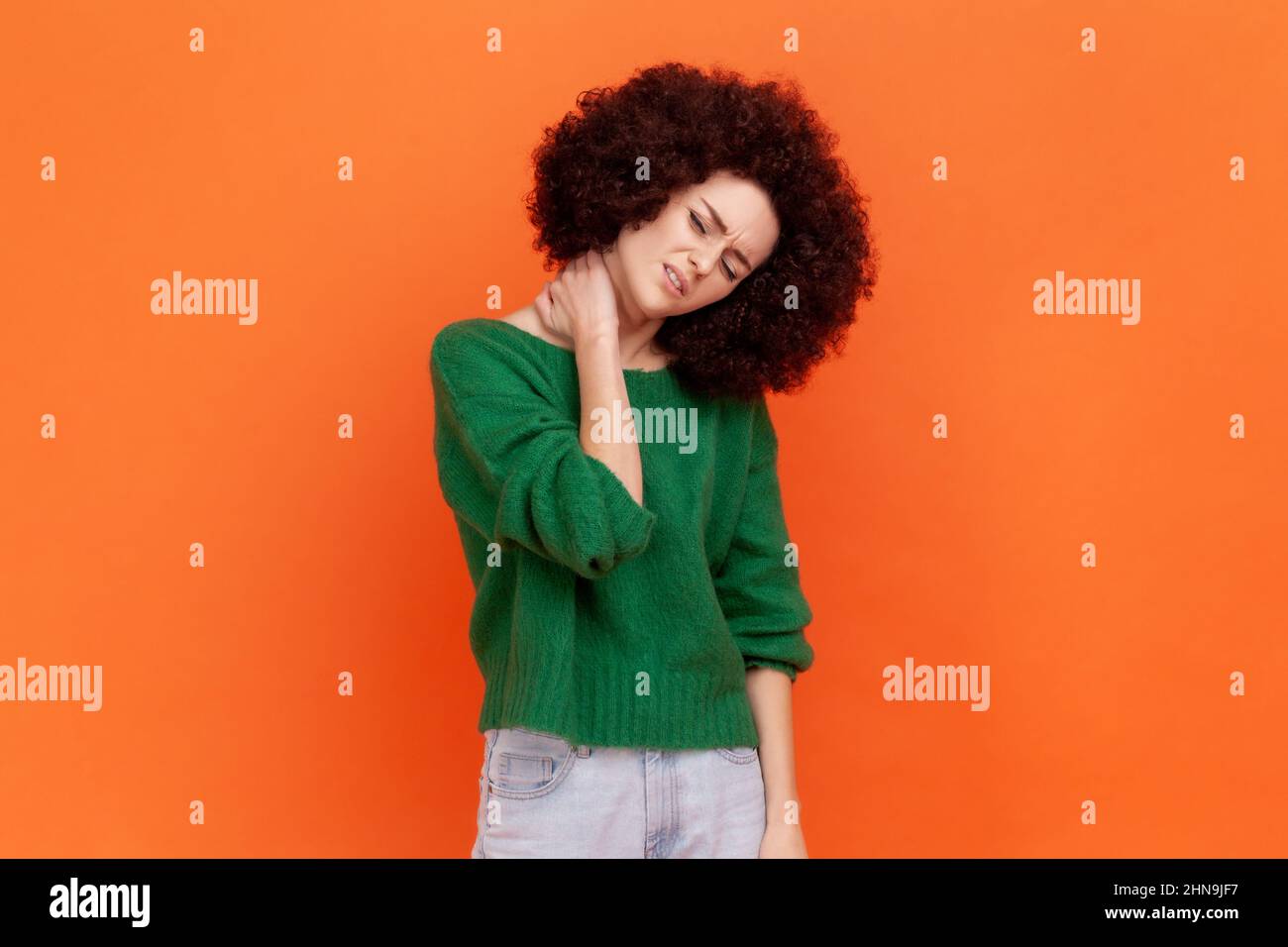 Problemi di salute. Ritratto di donna ammalata con acconciatura Afro indossare maglione verde casual stile in piedi e tenendo il suo collo doloroso. Studio interno girato isolato su sfondo arancione. Foto Stock