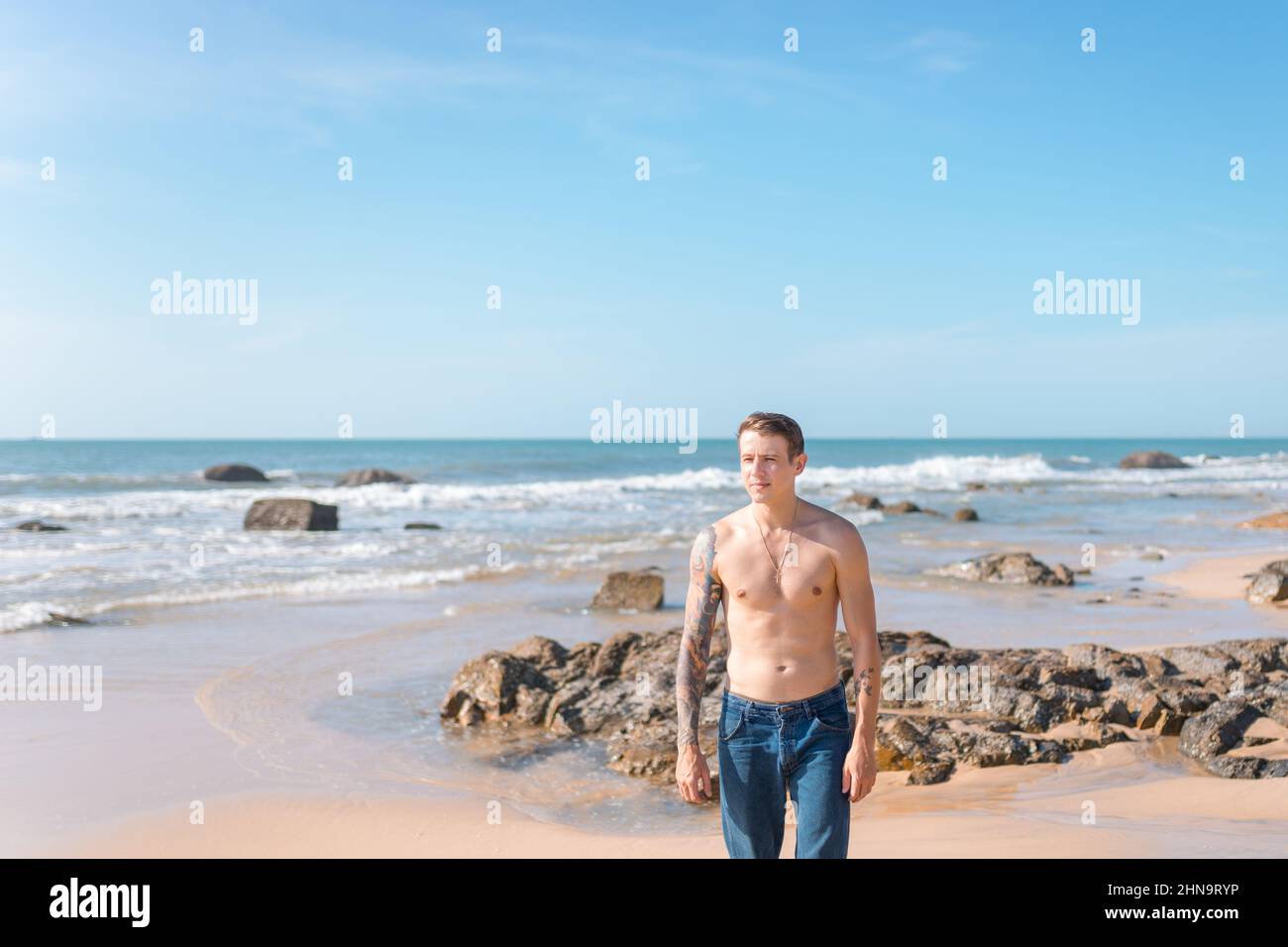 Attraente giovane caucasico nudo-chested uomo con braccio pieno tatuaggio che cammina sulla spiaggia a piedi nudi. Vista panoramica sull'oceano, foto mattutina, jeans. Foto Stock