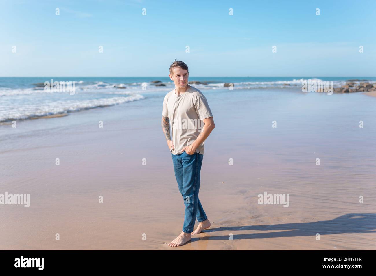 Attraente giovane uomo caucasico con tatuaggio a braccio pieno in abbigliamento casual camminando sulla spiaggia a piedi nudi. Vista panoramica sull'oceano, mattina. Foto di alta qualità Foto Stock