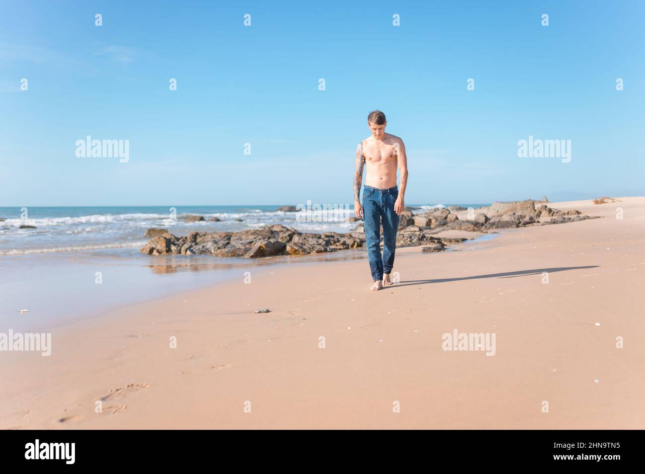 Attraente giovane caucasico nudo-chested uomo con braccio pieno tatuaggio che cammina sulla spiaggia a piedi nudi. Vista panoramica sull'oceano, foto mattutina, jeans. Foto Stock