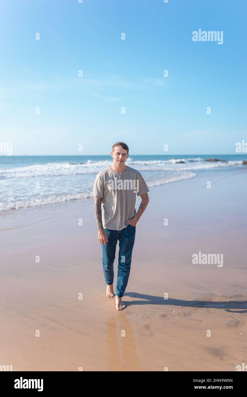 Attraente giovane uomo caucasico con tatuaggio a braccio pieno in abbigliamento casual camminando sulla spiaggia a piedi nudi. Vista panoramica sull'oceano, mattina. Foto di alta qualità Foto Stock