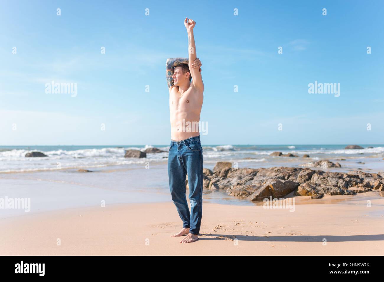 Bel vestito giovane caucasico nudo-vestito uomo con braccio pieno tatuaggio in piedi sulla spiaggia. Rocce, vista panoramica dell'oceano, che si allunga al mattino Foto Stock