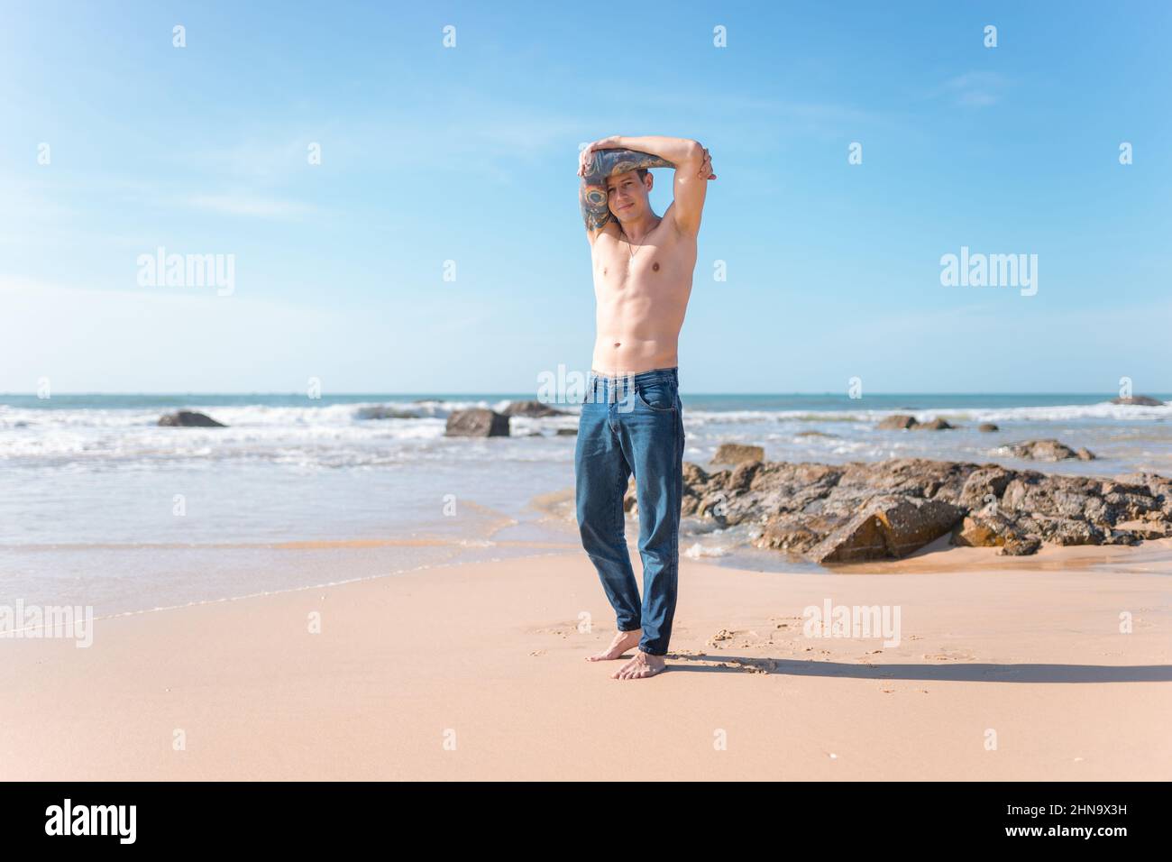 Bel vestito giovane caucasico nudo-vestito uomo con braccio pieno tatuaggio in piedi sulla spiaggia. Rocce, vista panoramica dell'oceano, che si allunga al mattino Foto Stock
