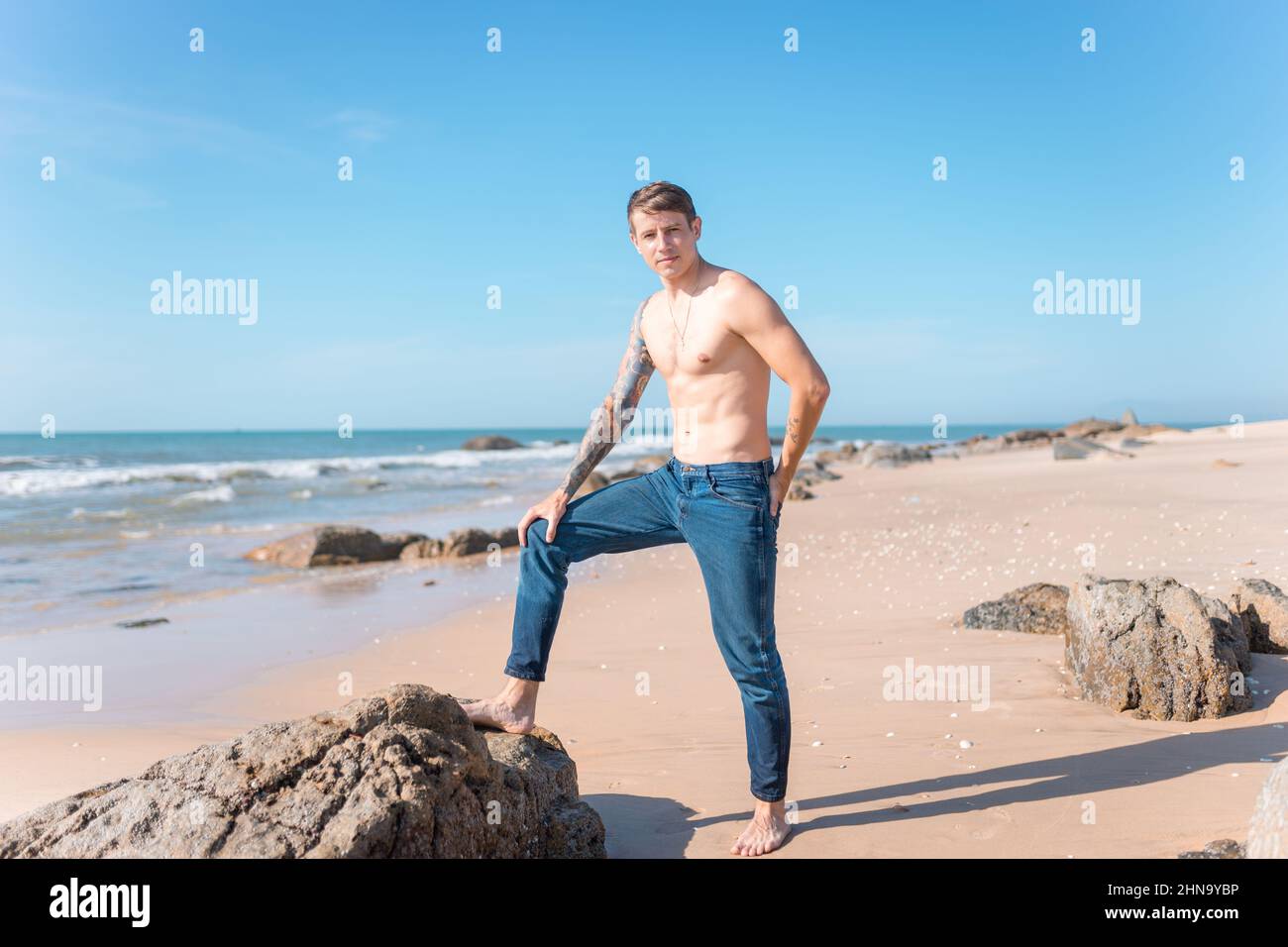 Attraente giovane uomo caucasico nudo-chested con tatuaggio full-ARM in piedi sulla spiaggia vicino alle rocce. Vista panoramica sull'oceano, mattina, con jeans. Foto Stock